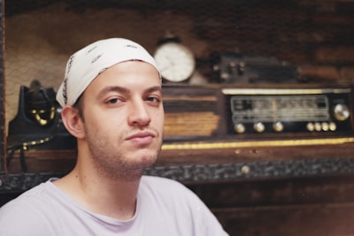 A man with a white bandana is sitting in front of a vintage radio and some kind of old equipment on shelves. The shelves have a rustic and industrial feel with wooden and metal elements.