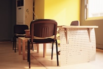 A small office space with a modern wooden desk and two chairs. The desk has a perforated metallic panel at the front, and the left chair is positioned partially under the table. An air conditioning unit is visible in the upper left corner. The room is well-lit with natural light coming through a window on the right, casting shadows on the floor.