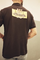 A person is standing with their back turned, wearing a black short-sleeved shirt featuring a design on the back. The design includes a drawing of a car with the words 'ART garage' above and below it. The person has visible tattoos on their arms and is wearing light blue jeans. The background is a neutral-toned, simple wall, enhancing the casual look of the scene.