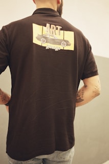 A person is standing with their back turned, wearing a black short-sleeved shirt featuring a design on the back. The design includes a drawing of a car with the words 'ART garage' above and below it. The person has visible tattoos on their arms and is wearing light blue jeans. The background is a neutral-toned, simple wall, enhancing the casual look of the scene.