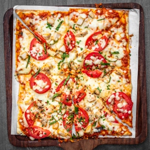A rectangular pizza with a crispy crust topped with slices of fresh tomatoes, scattered chicken pieces, melted mozzarella cheese, and garnished with green herbs. The pizza is served on a wooden tray with a sheet of parchment paper underneath.