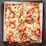 A rectangular pizza with a crispy crust topped with slices of fresh tomatoes, scattered chicken pieces, melted mozzarella cheese, and garnished with green herbs. The pizza is served on a wooden tray with a sheet of parchment paper underneath.