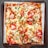 A rectangular pizza with a crispy crust topped with slices of fresh tomatoes, scattered chicken pieces, melted mozzarella cheese, and garnished with green herbs. The pizza is served on a wooden tray with a sheet of parchment paper underneath.
