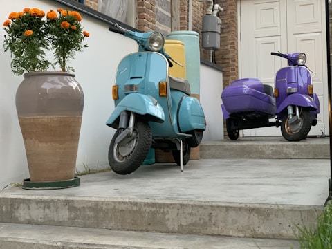 Two vintage scooters, one turquoise and the other purple with a sidecar, are parked on a concrete patio. A large potted plant with vibrant orange flowers stands next to the turquoise scooter. The setting includes a brick wall and a white door, contributing to an urban and stylish atmosphere.