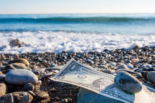 A one-dollar bill is partially covered by smooth pebbles on a rocky beach with ocean waves gently crashing in the background. The sunlight casts a bright and natural illumination on the scene.