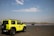 A bright yellow vehicle with a spare tire on the back is parked on a large open asphalt area. Orange traffic cones are scattered around the area, and there is a stack of tires in the background. The sky is partly cloudy, and the horizon shows a flat landscape with some vegetation.