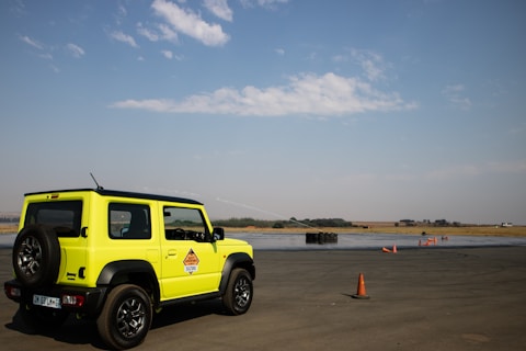 A bright yellow vehicle with a spare tire on the back is parked on a large open asphalt area. Orange traffic cones are scattered around the area, and there is a stack of tires in the background. The sky is partly cloudy, and the horizon shows a flat landscape with some vegetation.