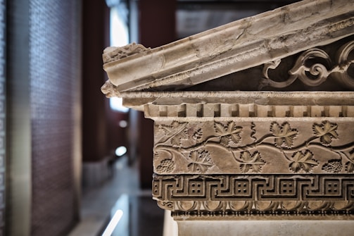 A detailed view of an intricately carved stone artifact, featuring classical architectural elements such as scrolls and floral motifs. The stone appears aged, with a subtle play of shadows and highlights. The background is softly blurred, suggesting an indoor setting possibly in a museum or historical site.