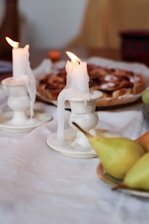 Two partially melted candles sit on white holders with visible wax drippings. They are placed on a white tablecloth along with a plate of pears and a dessert in the background, possibly a pie. The setting suggests a cozy, intimate atmosphere.