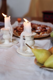 Two partially melted candles sit on white holders with visible wax drippings. They are placed on a white tablecloth along with a plate of pears and a dessert in the background, possibly a pie. The setting suggests a cozy, intimate atmosphere.