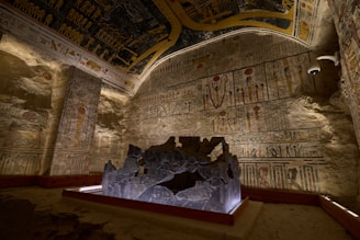 An ancient Egyptian tomb interior with walls and ceilings covered in detailed hieroglyphics and symbolic art. The area is dimly lit, highlighting the intricate carvings and colorful illustrations. A stone altar or centerpiece is positioned in the middle, surrounded by protective wooden barriers.