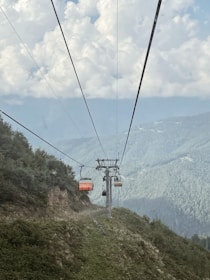 Cable cars travel along wires over a mountainous landscape with lush greenery. The sky is filled with fluffy clouds, providing a backdrop to the scene.