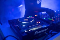 A dimly lit DJ setup featuring a close-up view of turntables and a mixer with various blinking lights. A partial view of a DJ's arm is seen adjusting the controls, surrounded by a blue ambient glow and a slightly smoky atmosphere.