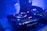 A dimly lit DJ setup featuring a close-up view of turntables and a mixer with various blinking lights. A partial view of a DJ's arm is seen adjusting the controls, surrounded by a blue ambient glow and a slightly smoky atmosphere.