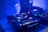 A dimly lit DJ setup featuring a close-up view of turntables and a mixer with various blinking lights. A partial view of a DJ's arm is seen adjusting the controls, surrounded by a blue ambient glow and a slightly smoky atmosphere.