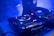 A dimly lit DJ setup featuring a close-up view of turntables and a mixer with various blinking lights. A partial view of a DJ's arm is seen adjusting the controls, surrounded by a blue ambient glow and a slightly smoky atmosphere.