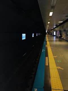 A dimly lit subway platform with a dark track on the left side and a well-lit waiting area on the right. Illuminated signage is visible on the wall above the tracks. The platform is marked with vibrant yellow tactile paving and directional arrows, with seating and advertisements further down the corridor.