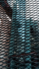 A close-up view of a metal mesh pattern, with crisscrossed wires creating a grid-like design. The background features blurred elements, suggesting an industrial or urban setting with hints of geometric structures.