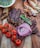 A wooden surface holds a gourmet spread including sliced, grilled steak garnished with chimichurri sauce, roasted cherry tomatoes on the vine, raw red onion rings, and grilled prawns with herbs. A bowl of chimichurri sauce is partially visible.