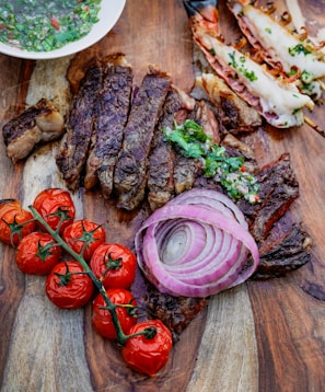 A wooden surface holds a gourmet spread including sliced, grilled steak garnished with chimichurri sauce, roasted cherry tomatoes on the vine, raw red onion rings, and grilled prawns with herbs. A bowl of chimichurri sauce is partially visible.
