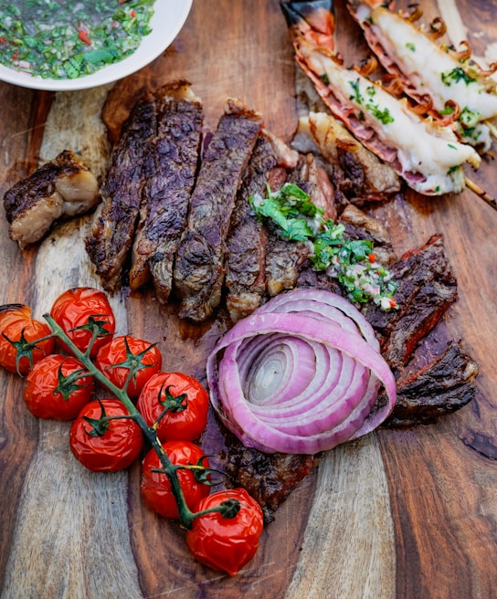 A wooden surface holds a gourmet spread including sliced, grilled steak garnished with chimichurri sauce, roasted cherry tomatoes on the vine, raw red onion rings, and grilled prawns with herbs. A bowl of chimichurri sauce is partially visible.
