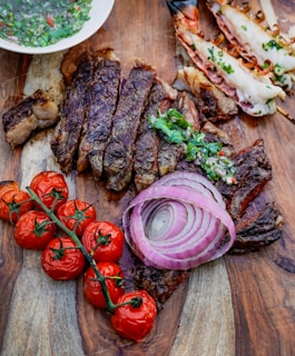 A wooden surface holds a gourmet spread including sliced, grilled steak garnished with chimichurri sauce, roasted cherry tomatoes on the vine, raw red onion rings, and grilled prawns with herbs. A bowl of chimichurri sauce is partially visible.