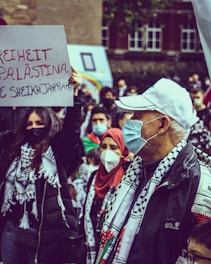 A group of people are gathered in a protest, wearing masks and holding signs. Several individuals wear scarves with black and white patterns, and a woman holds a sign with the message 'Freiheit Palästina' written in red and black. The background shows blurred buildings and more people.