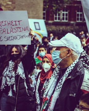 A group of people are gathered in a protest, wearing masks and holding signs. Several individuals wear scarves with black and white patterns, and a woman holds a sign with the message 'Freiheit Palästina' written in red and black. The background shows blurred buildings and more people.