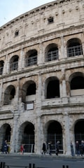 A close-up view of an ancient Roman amphitheater with multiple arches and weathered stone. The facade shows signs of aging and restoration, with visible scaffolding in some areas. Several people are walking along the sidewalk in front of the structure.