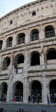 A close-up view of an ancient Roman amphitheater with multiple arches and weathered stone. The facade shows signs of aging and restoration, with visible scaffolding in some areas. Several people are walking along the sidewalk in front of the structure.