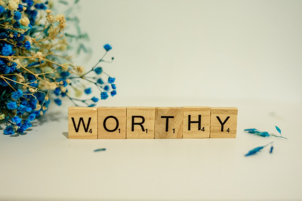 a wooden block spelling the word worthy next to a bouquet of blue flowers