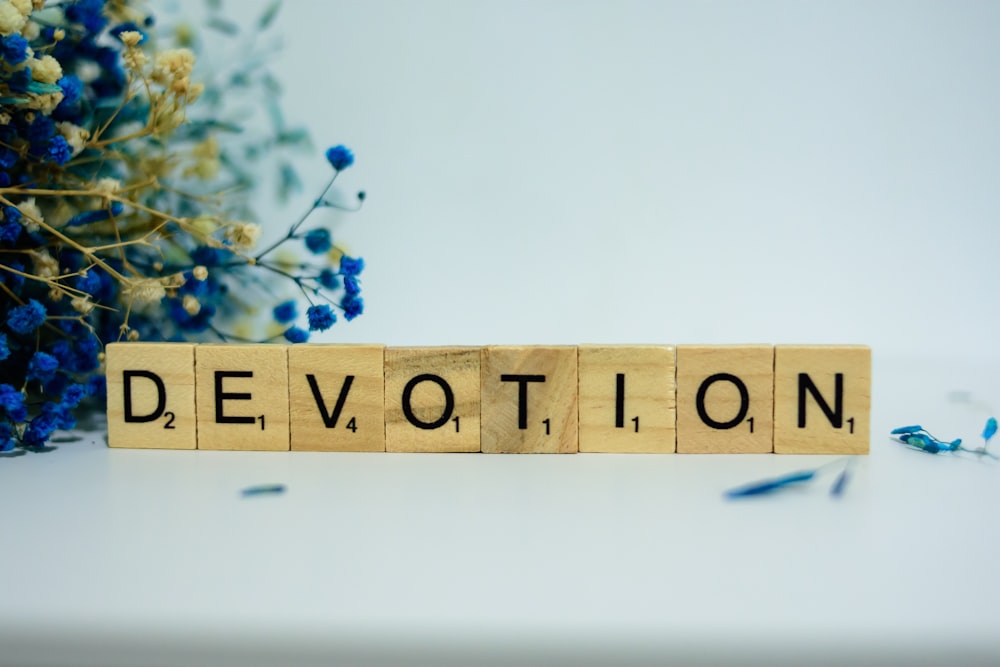 a wooden block spelling the word devotion next to blue flowers