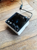 A compact audio device with multiple black knobs and a display panel sits on a wooden surface. A connected cable extends from the back, hinting at functionality as an audio interface or effects pedal.