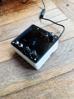 A compact audio device with multiple black knobs and a display panel sits on a wooden surface. A connected cable extends from the back, hinting at functionality as an audio interface or effects pedal.