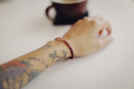 A tattooed arm with a red beaded bracelet rests on a white surface. In the background, there is a dark-colored cup and saucer, slightly out of focus.