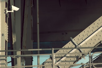 An industrial structure featuring metal beams and a security camera mounted on one side. The beams are reinforced with bolts and have a weathered appearance. The lighting casts shadows across the structure, creating a contrast between light and dark areas. The background has numbers '75' and '74' painted on the wall.