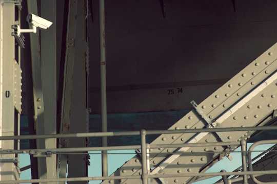 An industrial structure featuring metal beams and a security camera mounted on one side. The beams are reinforced with bolts and have a weathered appearance. The lighting casts shadows across the structure, creating a contrast between light and dark areas. The background has numbers '75' and '74' painted on the wall.