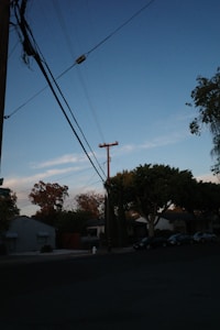 A residential street scene during the early evening with a clear sky. Telephone poles and power lines cross overhead. Trees and parked cars line the street, with small houses visible in the background. The sky has a gradient from a deep blue to a light blue near the horizon.