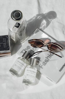 A glass jar and a deck of cards are placed on a white surface alongside two perfume bottles labeled 'Osmanthus 16' and 'Blackcurrant'. A pair of tortoiseshell sunglasses rests on a fashion magazine.