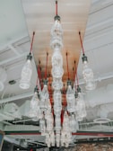 Multiple glass bottles hang upside down from a ceiling, with each bottle having a light bulb at the base. They are suspended from red cords, creating an artistic chandelier effect. The ceiling features industrial elements such as exposed pipes and vents.