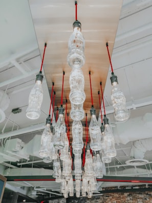Multiple glass bottles hang upside down from a ceiling, with each bottle having a light bulb at the base. They are suspended from red cords, creating an artistic chandelier effect. The ceiling features industrial elements such as exposed pipes and vents.