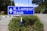 A blue road sign displaying directions to K. Lumpur and Raub stands prominently against a background of greenery and a clear sky. The sign is surrounded by small bushes with small yellow flowers, and the road is visible in the background.