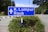 A blue road sign displaying directions to K. Lumpur and Raub stands prominently against a background of greenery and a clear sky. The sign is surrounded by small bushes with small yellow flowers, and the road is visible in the background.