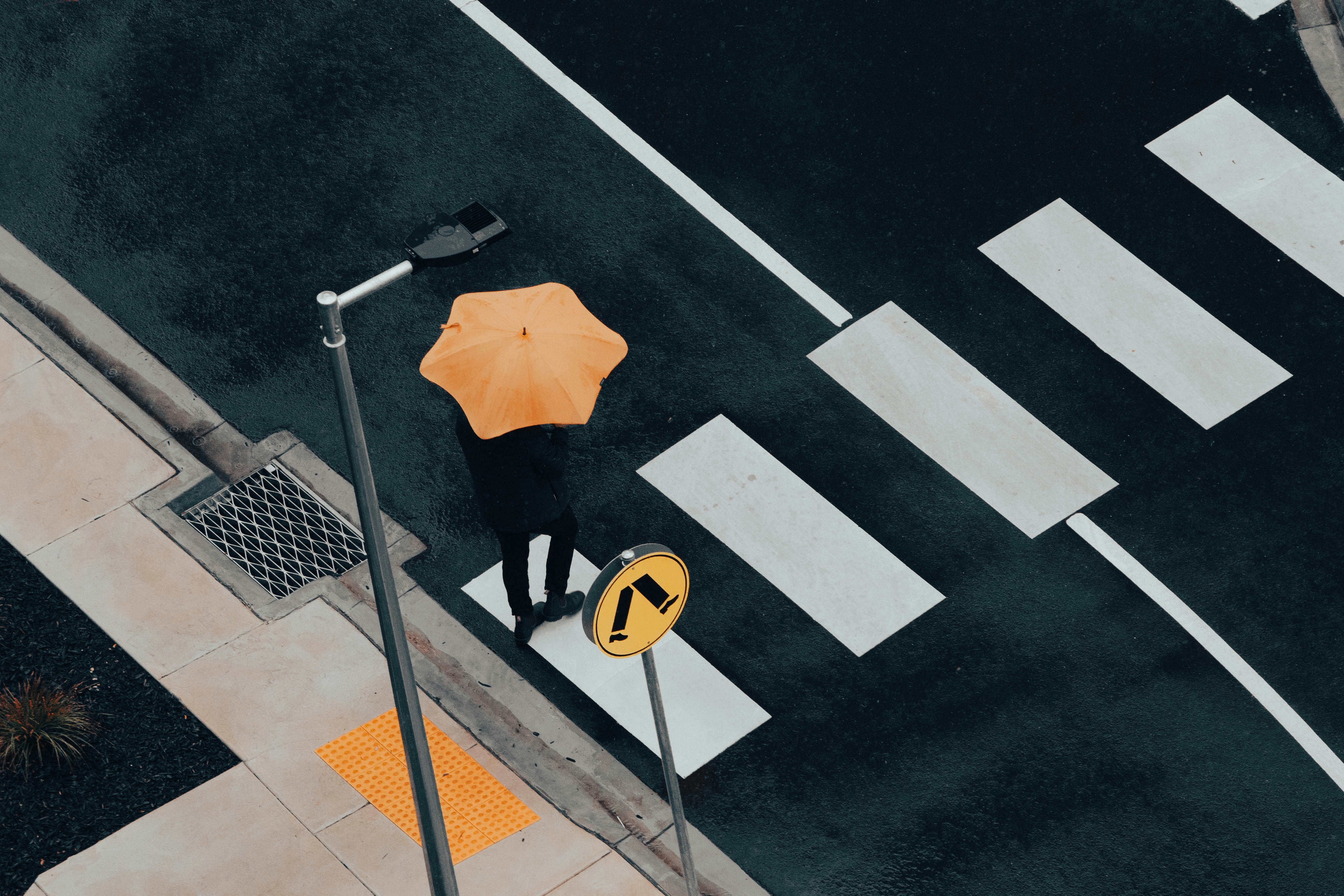 A man crossing a road at a road crossing during a rainy day. The man holds an unbrella.