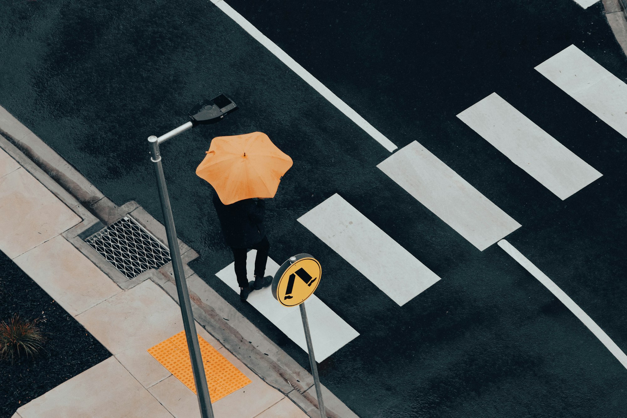 A man crossing a road at a road crossing during a rainy day. The man holds an unbrella.