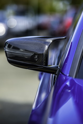 A close-up view of a carbon fiber side mirror on a sleek, high-performance sports car. The side mirror features a modern aerodynamic design, indicating its likely use in a fast vehicle. The car is painted in a vivid blue color, and the background is blurred, creating a bokeh effect that emphasizes the subject.