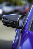 A close-up view of a carbon fiber side mirror on a sleek, high-performance sports car. The side mirror features a modern aerodynamic design, indicating its likely use in a fast vehicle. The car is painted in a vivid blue color, and the background is blurred, creating a bokeh effect that emphasizes the subject.