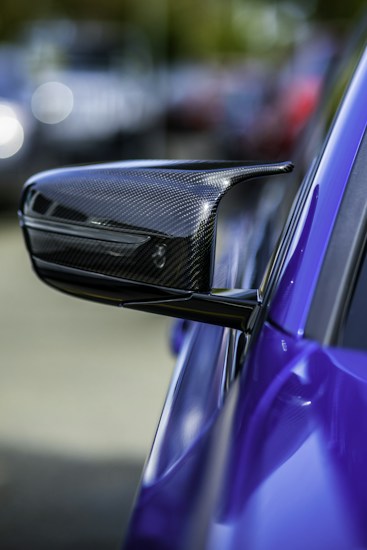 A close-up view of a carbon fiber side mirror on a sleek, high-performance sports car. The side mirror features a modern aerodynamic design, indicating its likely use in a fast vehicle. The car is painted in a vivid blue color, and the background is blurred, creating a bokeh effect that emphasizes the subject.