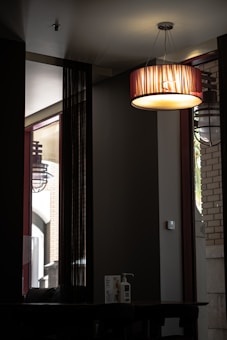 A dimly lit interior space showcasing a modern, cylindrical ceiling light fixture that emits a warm glow. The walls are dark, with large windows allowing some natural light to enter. A table beneath the light features a bottle of hand sanitizer and a small sign. Vertical black curtains or panels hang by the windows.