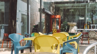A courtyard with a variety of geometric chairs in vibrant colors such as yellow, blue, and red arranged around tables. There's a red phone booth in the background against a glass and brick building facade. Decorative elements, including a statue and a sculptural fountain, add an artistic touch to the setting.